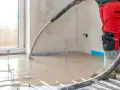 Installazione riscaldamento a pavimento: un operaio in tuta rossa versa il massetto su tubi e isolante.