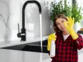 Donna con guanti gialli e spray, fa ok vicino a rubinetti neri che non si rovinano, mostrando la pulizia della cucina.