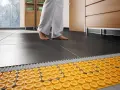 Piedi nudi su pavimento scuro, con sistema di riscaldamento a pavimento su pavimento esistente visibile sotto.