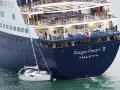Una piccola barca a vela si trova in una **posa a tolda di nave irregolare** accanto alla massiccia nave da crociera "Saga Pearl II".
