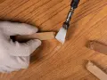 Mano guantata rimuove residui di colla da un parquet in legno. Un attrezzo metallico raschia via il materiale appiccicoso, mentre altri pezzi di colla sono visibili sul pavimento.