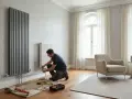 Un tecnico esegue il montaggio termosifoni in un soggiorno moderno. Attrezzi a terra, un radiatore verticale grigio e uno bianco.