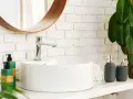 Lavabo bianco moderno con rubinetto cromato, fissato con silicone. Sul ripiano, dispenser sapone, porta spazzolino e piante verdi.