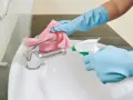 Mani guantate spruzzano detergente su un rubinetto del bagno, pronto per essere pulito con un panno rosa.