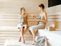 Due donne sorridono e chiacchierano in una sauna finlandese, avvolte in asciugamani.