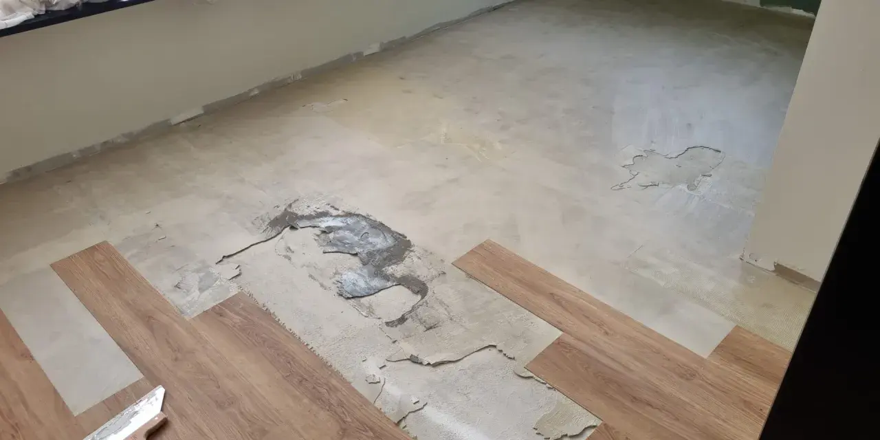 Il pavimento in PVC si solleva in alcune aree, mostrando il sottofondo danneggiato. Si vedono nuove doghe in legno e una spatola.