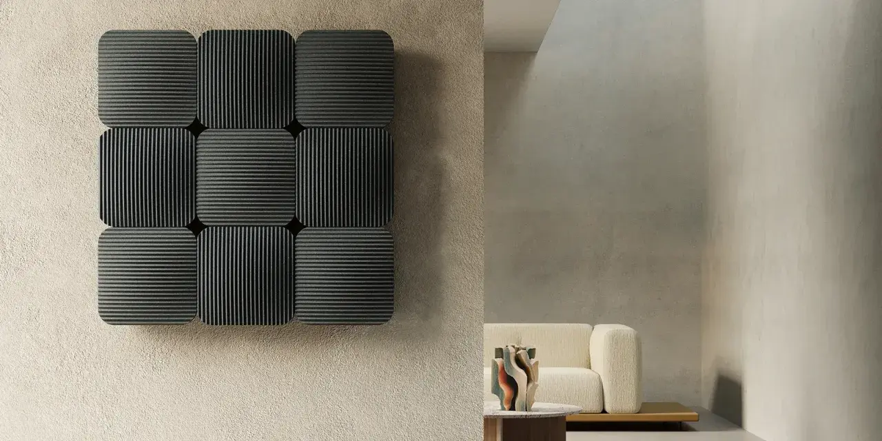 Pannelli neri con righe verticali e orizzontali, che ricordano la convezione termosifone, su una parete grezza. In secondo piano, un divano beige e un tavolino.