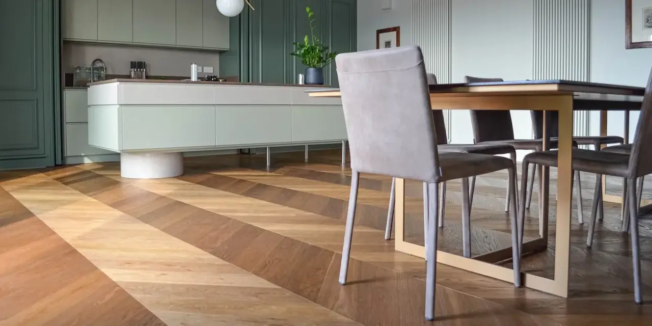 Un elegante parquet a spina di pesce crea un disegno dinamico sotto un tavolo da pranzo e una moderna cucina verde.