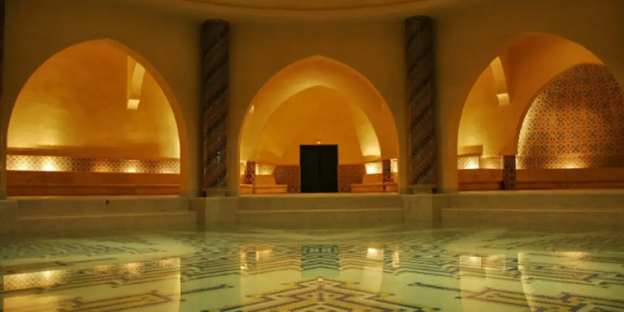 Interno di un bagno dei turchi con piscina e mosaici geometrici. Archi moreschi e colonne decorate creano un'atmosfera orientale.