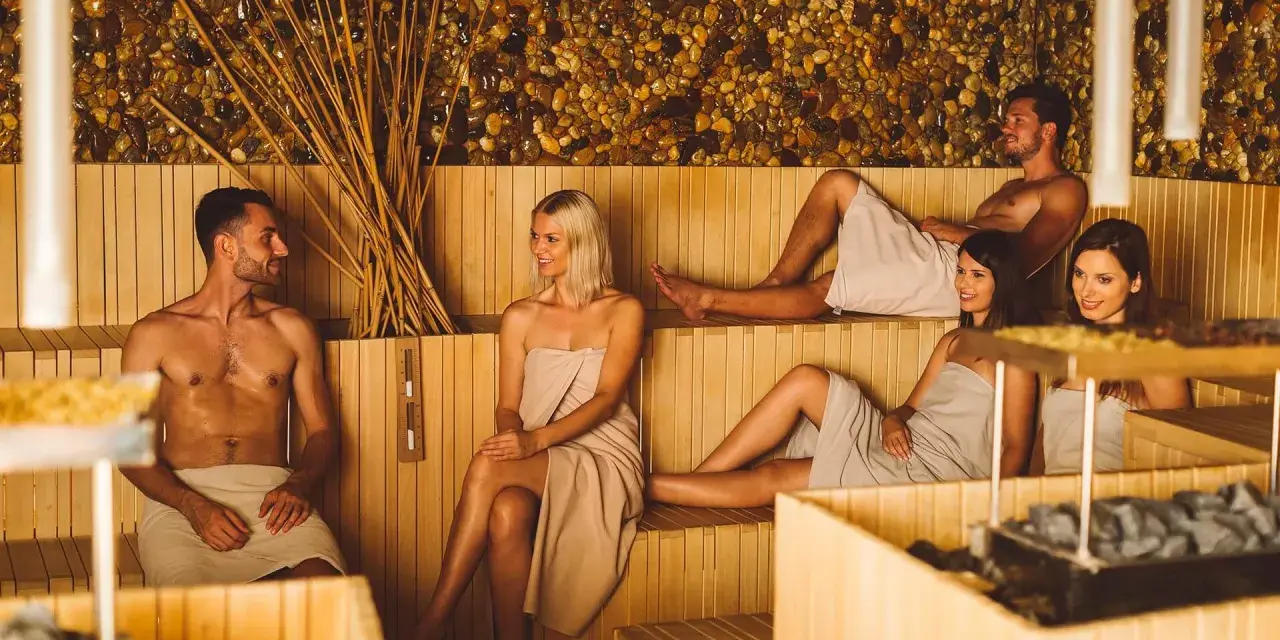 Un gruppo di persone nudi in sauna, avvolti in asciugamani, chiacchiera rilassato.