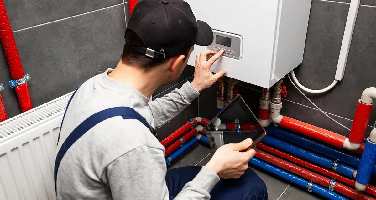 Uomo con tablet e cappellino esegue pulizia impianto riscaldamento fai da te, controllando la caldaia e i tubi colorati.