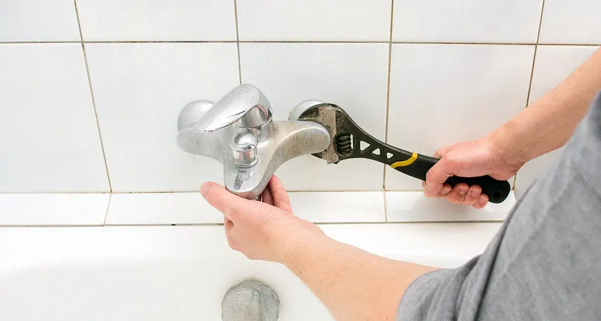 Mano con chiave inglese ripara un rubinetto che perde dal miscelatore, sopra la vasca da bagno.