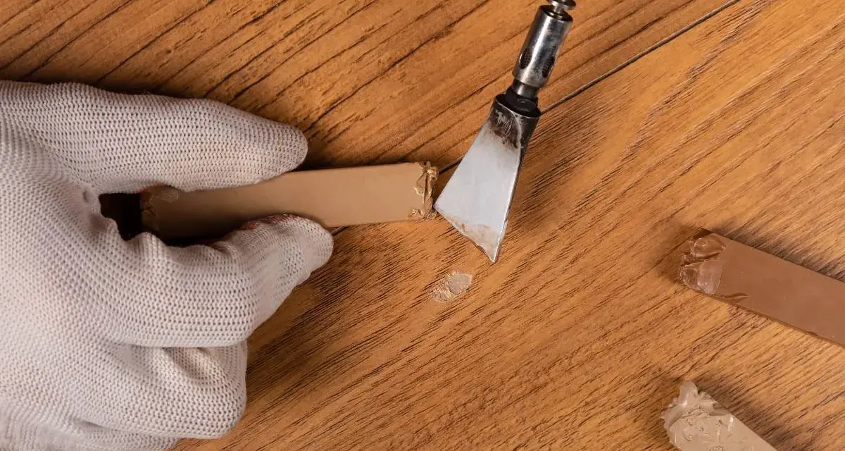Mano guantata rimuove residui di colla da un parquet in legno. Un attrezzo metallico raschia via il materiale appiccicoso, mentre altri pezzi di colla sono visibili sul pavimento.