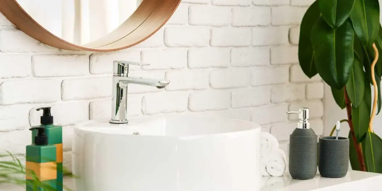 Lavabo bianco moderno con rubinetto cromato, fissato con silicone. Sul ripiano, dispenser sapone, porta spazzolino e piante verdi.
