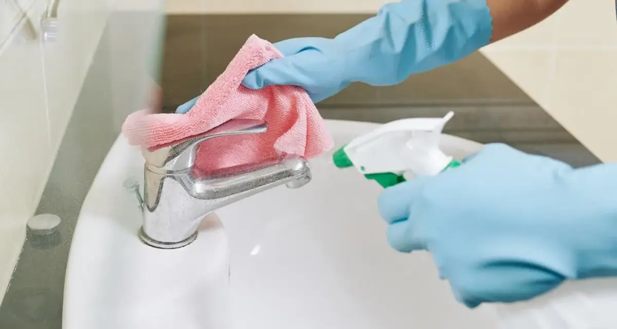 Mani guantate spruzzano detergente su un rubinetto del bagno, pronto per essere pulito con un panno rosa.