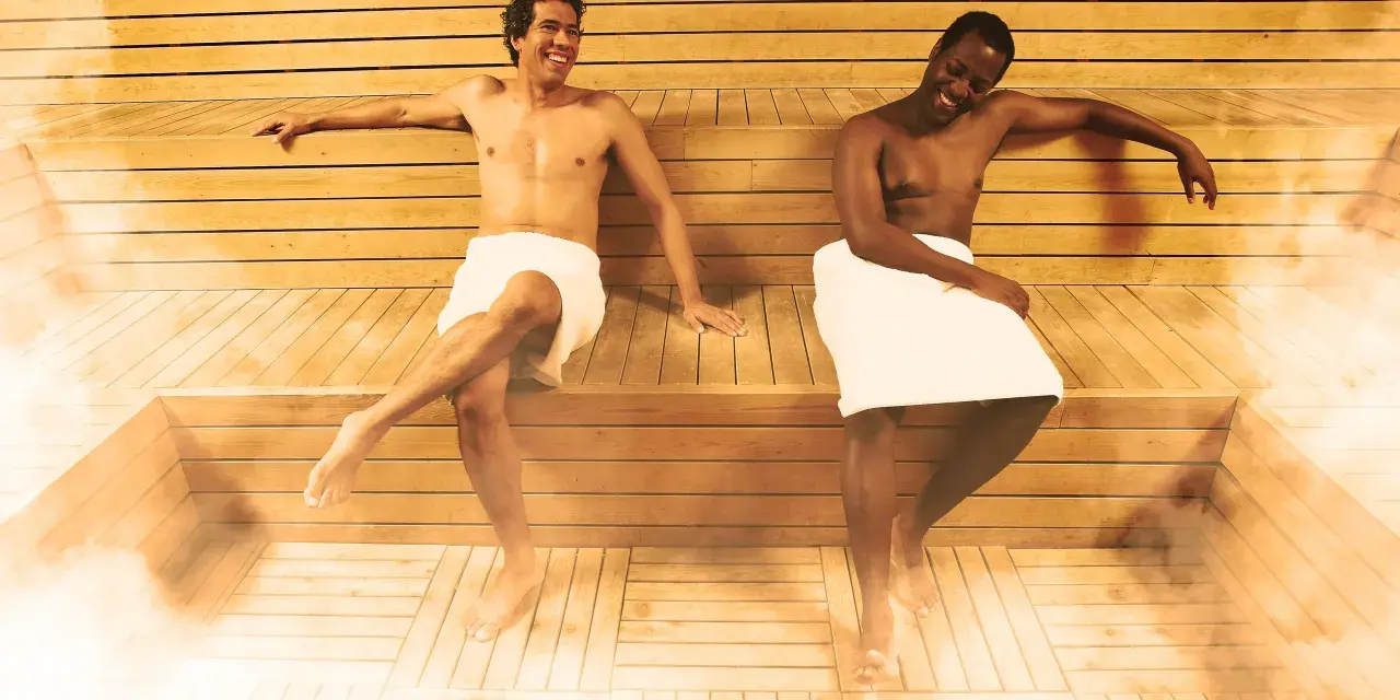 Due uomini sorridono nella sauna, godendosi il calore e il vapore. La sauna può offrire benefici per la pelle, inclusi quelli per la cellulite.