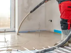 Installazione riscaldamento a pavimento: un operaio in tuta rossa versa il massetto su tubi e isolante.