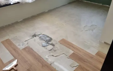 Il pavimento in PVC si solleva in alcune aree, mostrando il sottofondo danneggiato. Si vedono nuove doghe in legno e una spatola.
