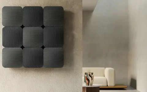 Pannelli neri con righe verticali e orizzontali, che ricordano la convezione termosifone, su una parete grezza. In secondo piano, un divano beige e un tavolino.