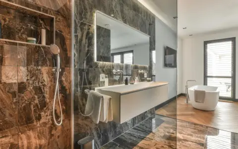 Bagno di lusso con doccia e vasca. Rubinetteria Bossini cromata, lavabo sospeso e specchio illuminato. Marmo scuro e parquet.