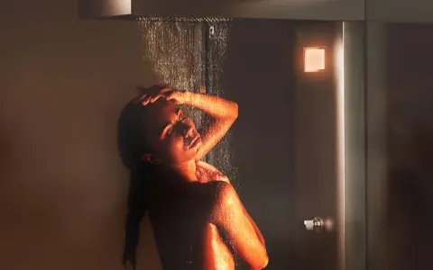 Donna sotto una doccia emozionale, illuminata da luci calde, con acqua che scorre sul suo corpo.