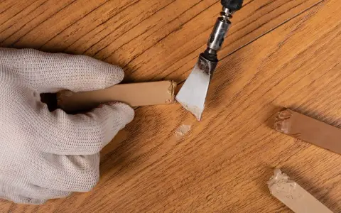 Mano guantata rimuove residui di colla da un parquet in legno. Un attrezzo metallico raschia via il materiale appiccicoso, mentre altri pezzi di colla sono visibili sul pavimento.