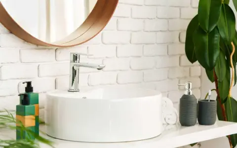 Lavabo bianco moderno con rubinetto cromato, fissato con silicone. Sul ripiano, dispenser sapone, porta spazzolino e piante verdi.