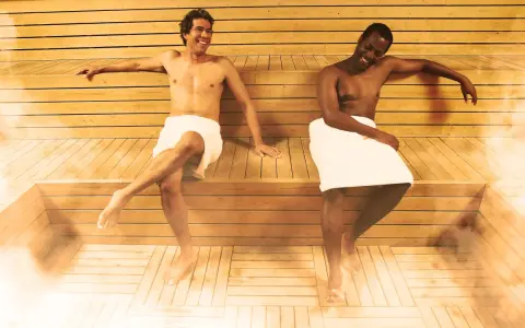 Due uomini sorridono nella sauna, godendosi il calore e il vapore. La sauna può offrire benefici per la pelle, inclusi quelli per la cellulite.