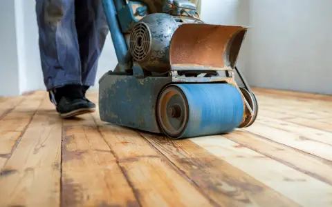 Levigatura parquet: un operaio usa una levigatrice per preparare il pavimento prima dell'imbiancatura. Il legno grezzo risplende sotto la macchina.
