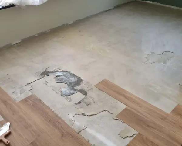 Il pavimento in PVC si solleva in alcune aree, mostrando il sottofondo danneggiato. Si vedono nuove doghe in legno e una spatola.