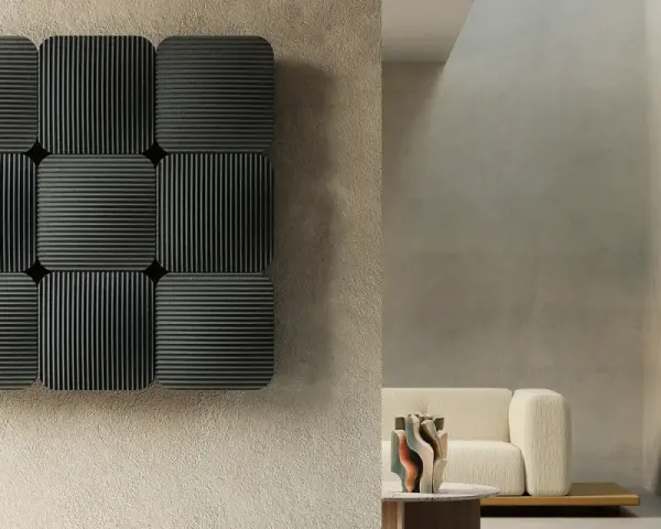 Pannelli neri con righe verticali e orizzontali, che ricordano la convezione termosifone, su una parete grezza. In secondo piano, un divano beige e un tavolino.