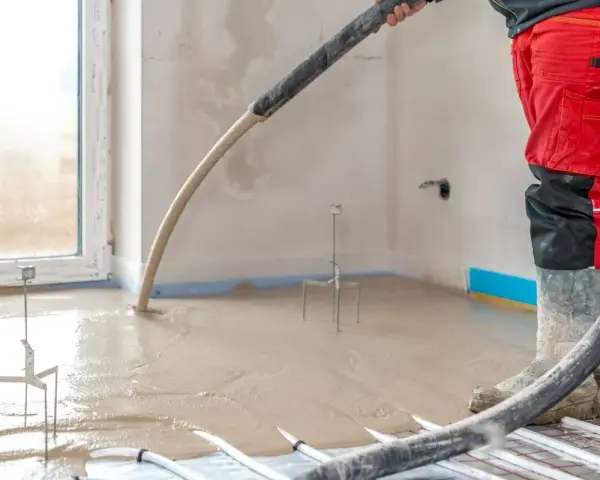Installazione riscaldamento a pavimento: un operaio in tuta rossa versa il massetto su tubi e isolante.