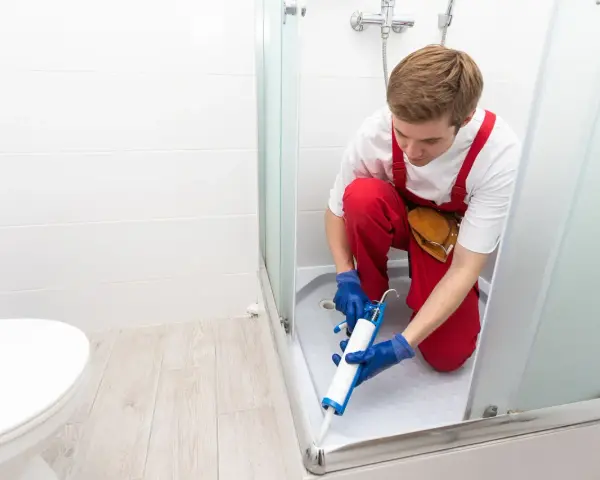 Un operaio con tuta rossa sigilla la fuga tra piatto doccia e piastrelle, prevenendo infiltrazioni d'acqua.