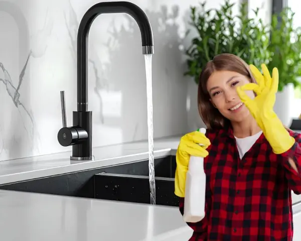 Donna con guanti gialli e spray, fa ok vicino a rubinetti neri che non si rovinano, mostrando la pulizia della cucina.