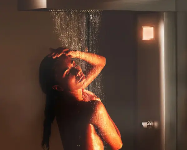 Donna sotto una doccia emozionale, illuminata da luci calde, con acqua che scorre sul suo corpo.