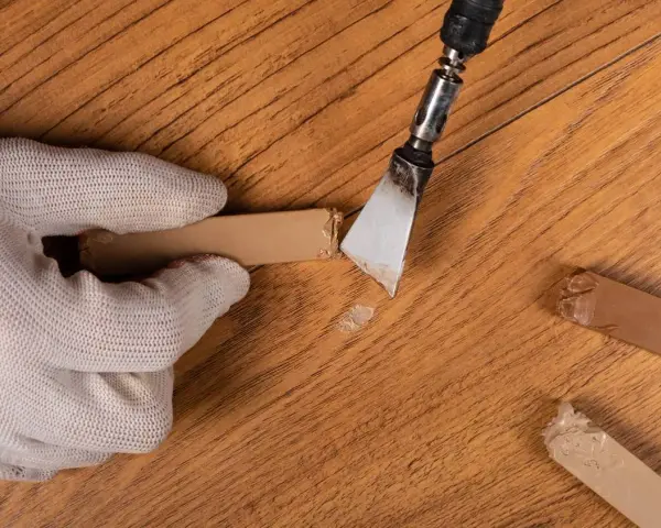 Mano guantata rimuove residui di colla da un parquet in legno. Un attrezzo metallico raschia via il materiale appiccicoso, mentre altri pezzi di colla sono visibili sul pavimento.