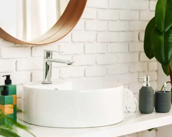 Lavabo bianco moderno con rubinetto cromato, fissato con silicone. Sul ripiano, dispenser sapone, porta spazzolino e piante verdi.