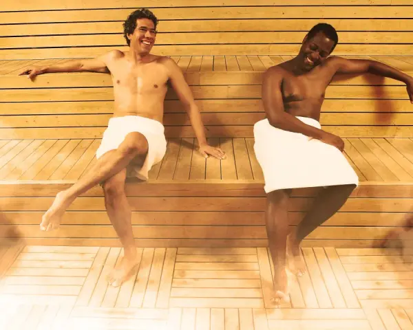 Due uomini sorridono nella sauna, godendosi il calore e il vapore. La sauna può offrire benefici per la pelle, inclusi quelli per la cellulite.