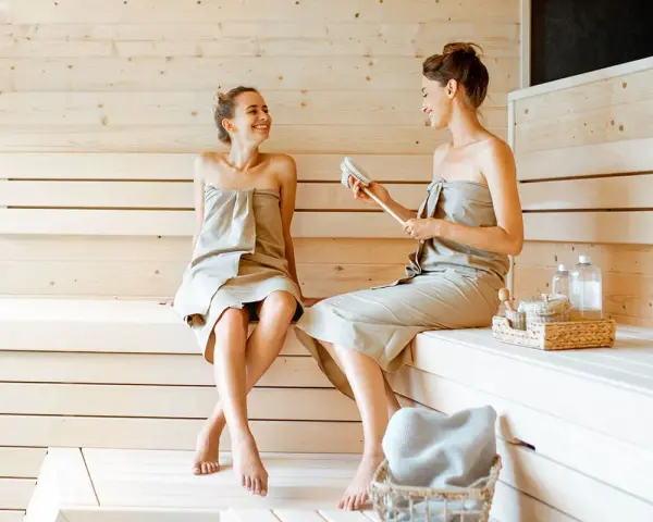 Due donne sorridono e chiacchierano in una sauna finlandese, avvolte in asciugamani.