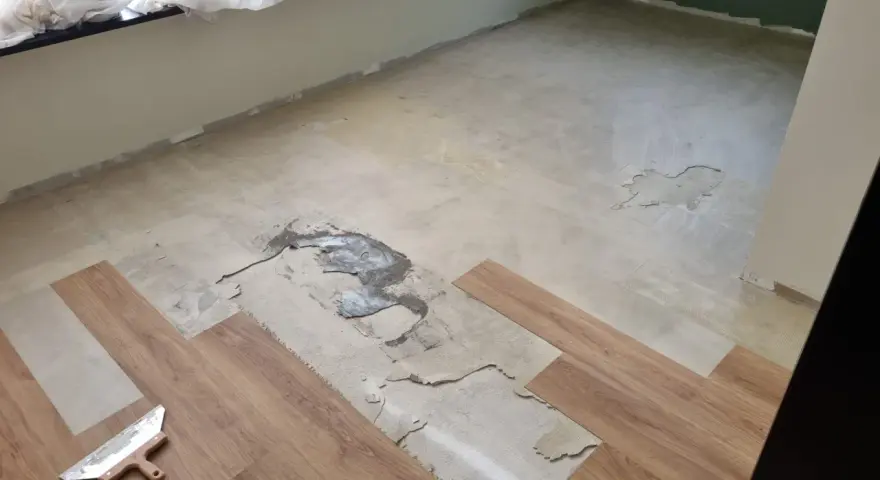 Il pavimento in PVC si solleva in alcune aree, mostrando il sottofondo danneggiato. Si vedono nuove doghe in legno e una spatola.