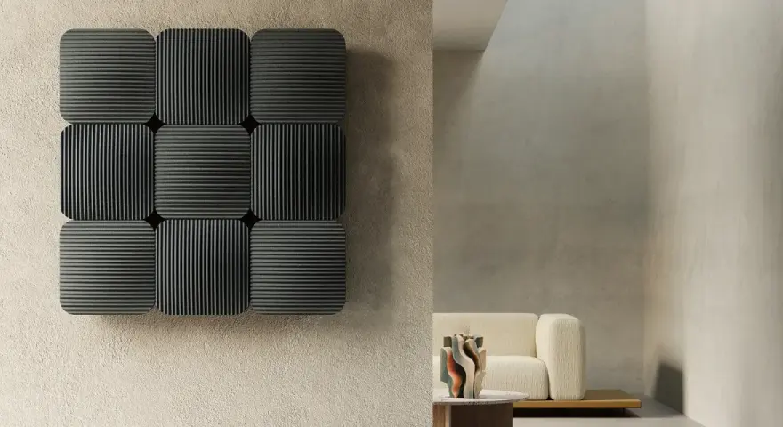 Pannelli neri con righe verticali e orizzontali, che ricordano la convezione termosifone, su una parete grezza. In secondo piano, un divano beige e un tavolino.