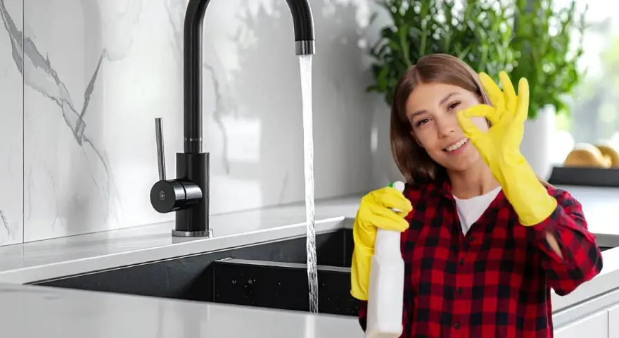 Donna con guanti gialli e spray, fa ok vicino a rubinetti neri che non si rovinano, mostrando la pulizia della cucina.