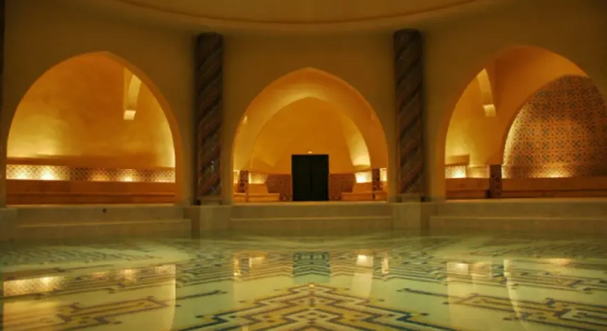 Interno di un bagno dei turchi con piscina e mosaici geometrici. Archi moreschi e colonne decorate creano un'atmosfera orientale.