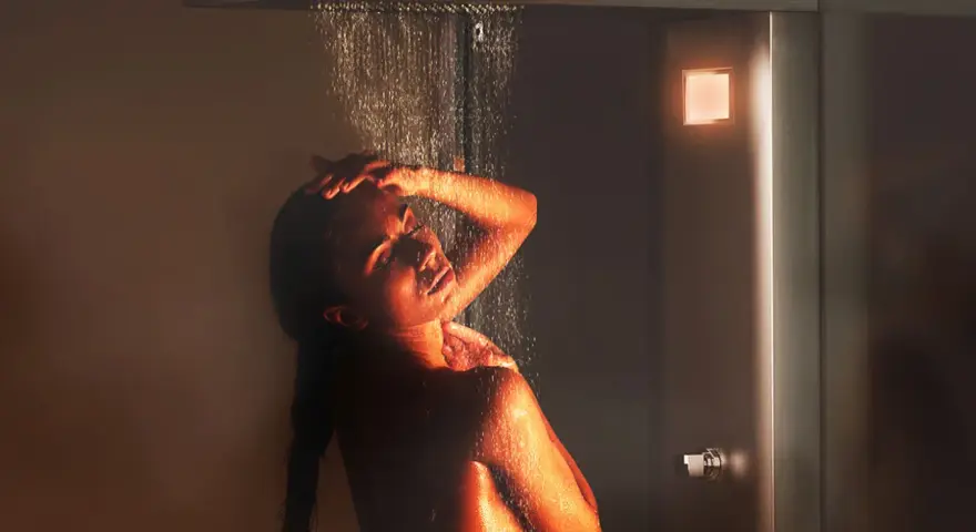 Donna sotto una doccia emozionale, illuminata da luci calde, con acqua che scorre sul suo corpo.