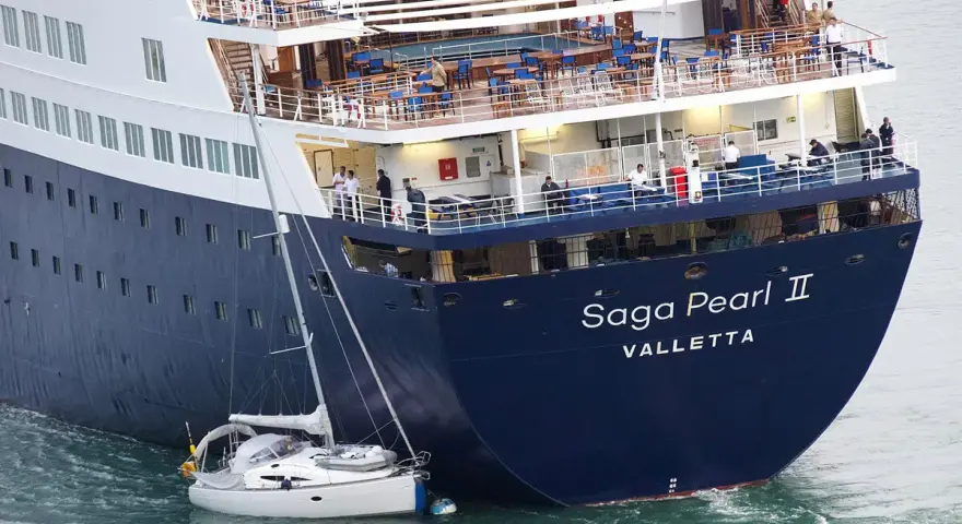 Una piccola barca a vela si trova in una **posa a tolda di nave irregolare** accanto alla massiccia nave da crociera "Saga Pearl II".
