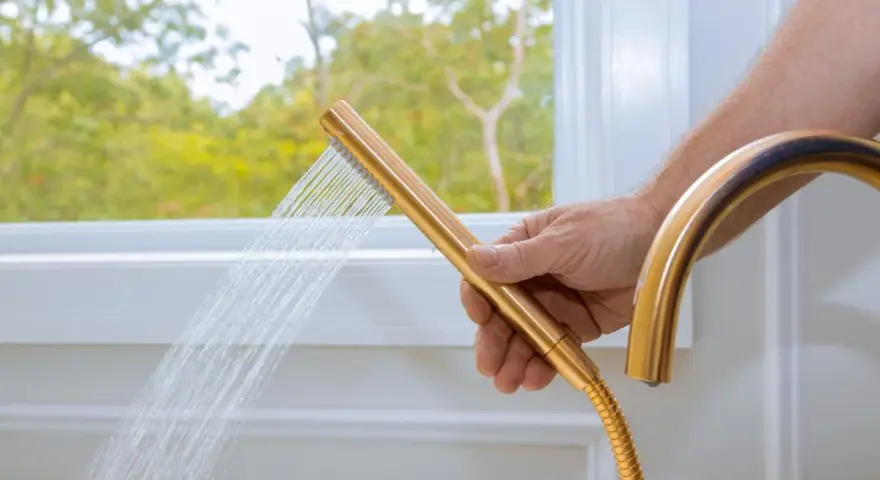 Mano che tiene un soffione doccia dorato, con acqua che scorre. Un esempio pratico su come pulire il tubo flessibile della doccia per un getto ottimale.