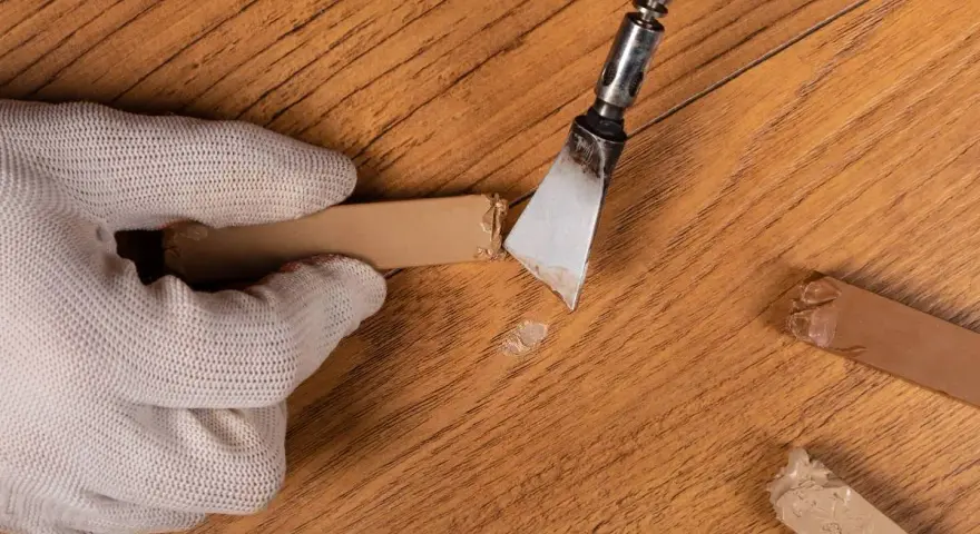 Mano guantata rimuove residui di colla da un parquet in legno. Un attrezzo metallico raschia via il materiale appiccicoso, mentre altri pezzi di colla sono visibili sul pavimento.