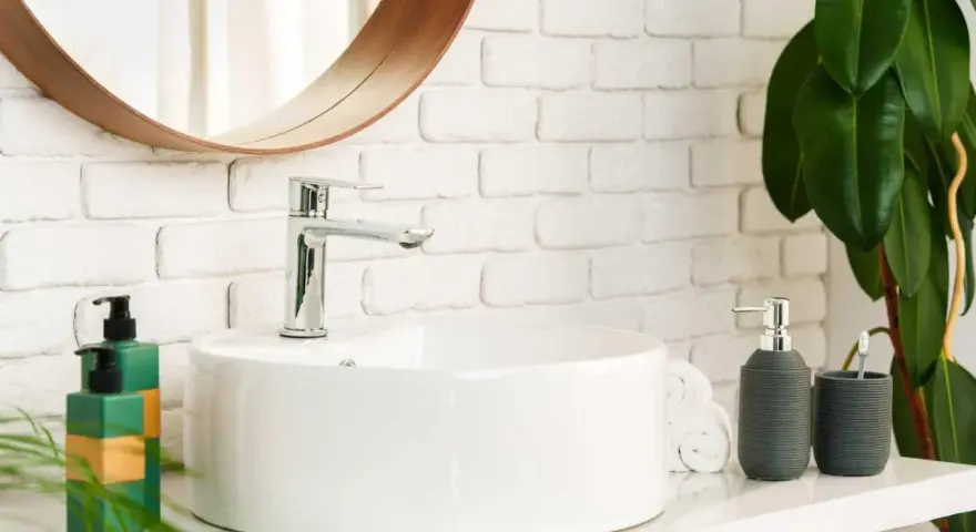 Lavabo bianco moderno con rubinetto cromato, fissato con silicone. Sul ripiano, dispenser sapone, porta spazzolino e piante verdi.