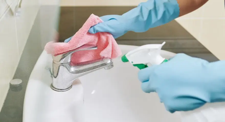 Mani guantate spruzzano detergente su un rubinetto del bagno, pronto per essere pulito con un panno rosa.