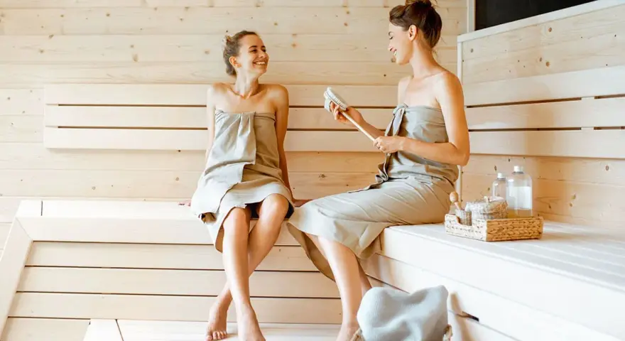 Due donne sorridono e chiacchierano in una sauna finlandese, avvolte in asciugamani.
